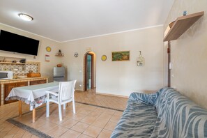 A living room with a sofa, a dining table with chairs, a kitchen area with a microwave, and a TV mounted on the wall.