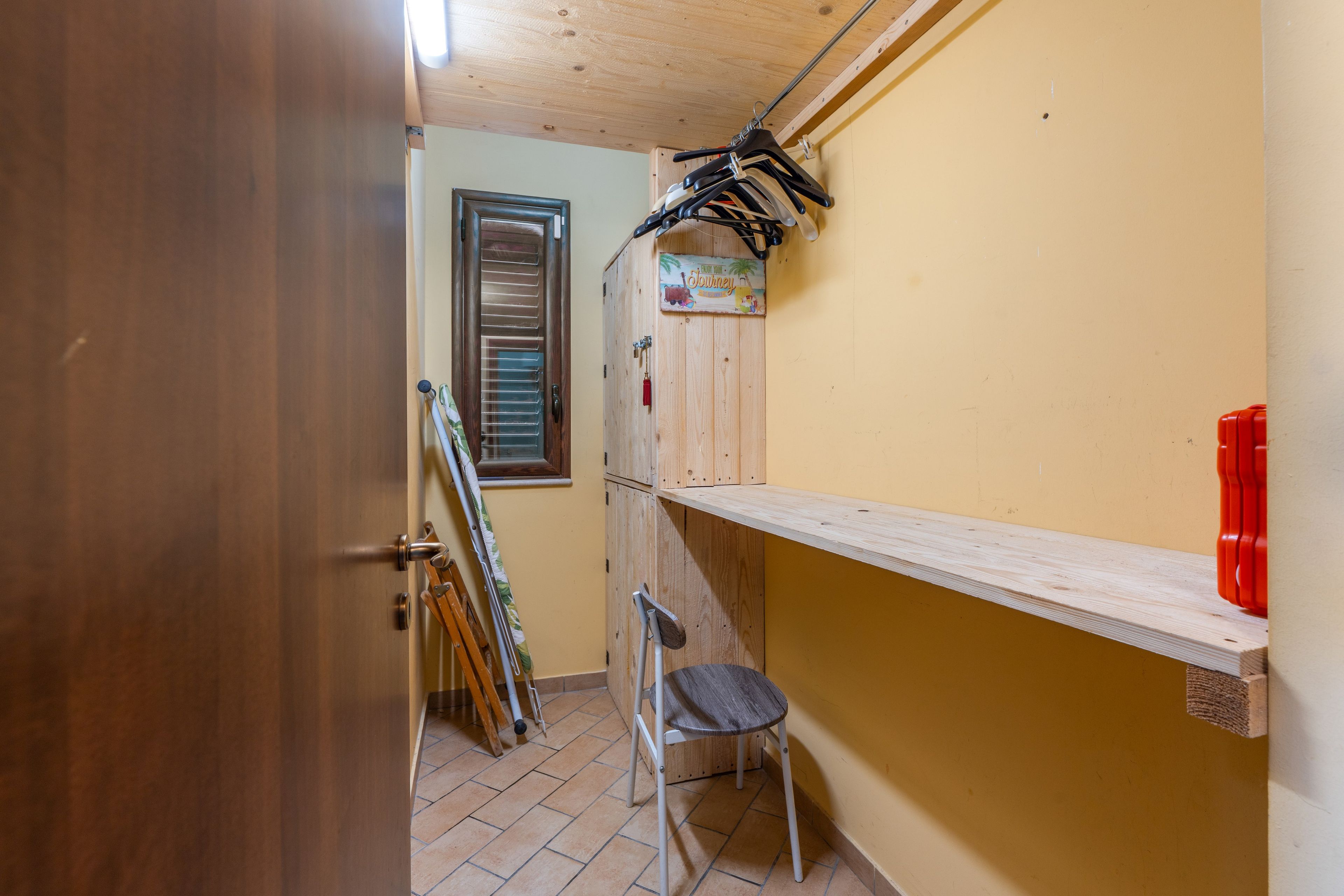 Ein schmaler Flur mit einer Holztür, einem Fenster mit Jalousien, einem Holzkasten mit dem Schriftzug 'Enjoy Your Journey', einem Stuhl und einem roten Gegenstand an der Wand.