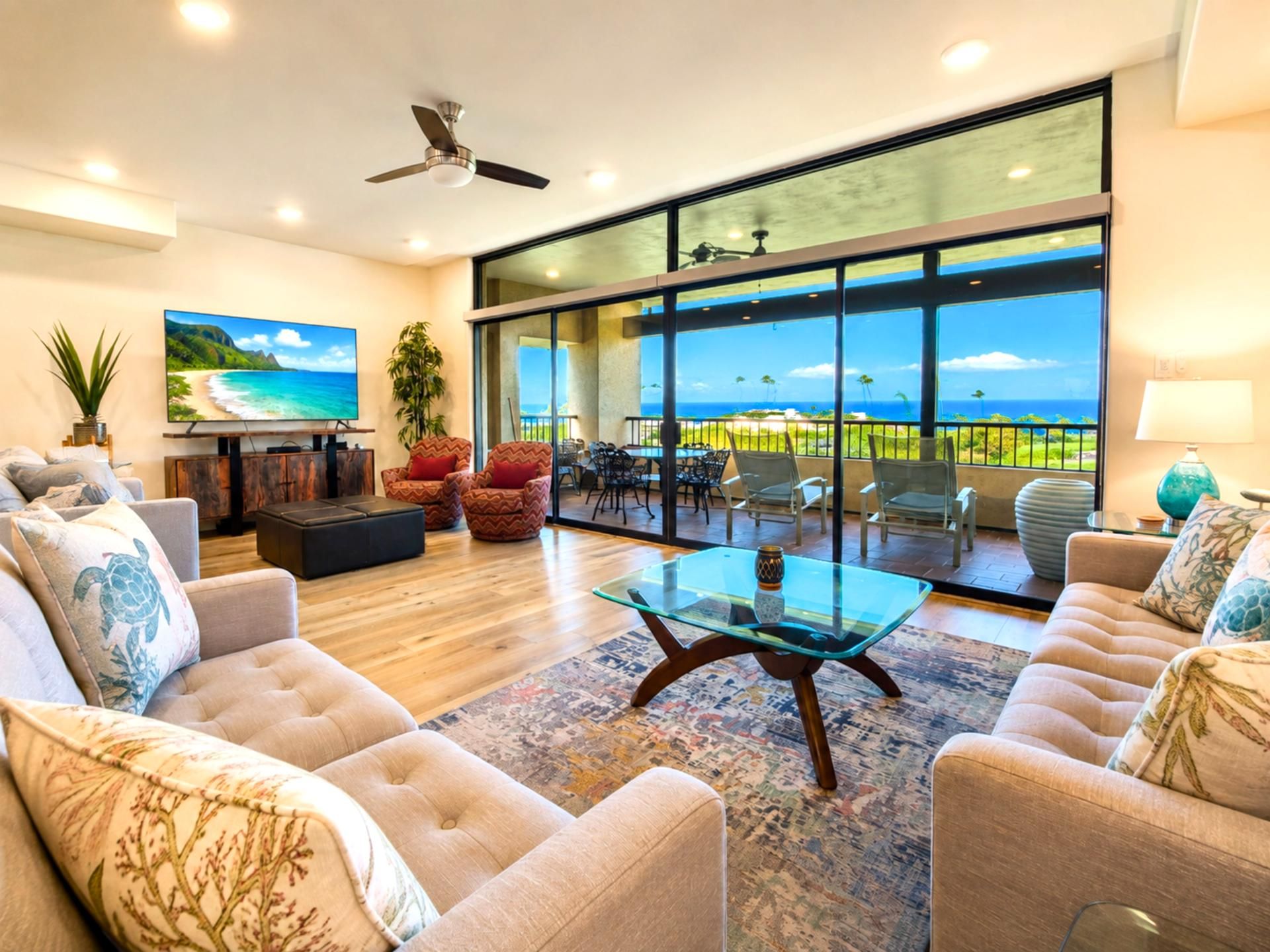 A spacious living room with a large TV, comfortable seating, a glass coffee table, and a balcony with a view of the ocean.