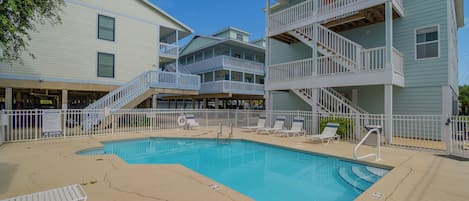 A pool area with a clear blue pool, white lounge chairs, and a staircase leading to a multi-level building.
