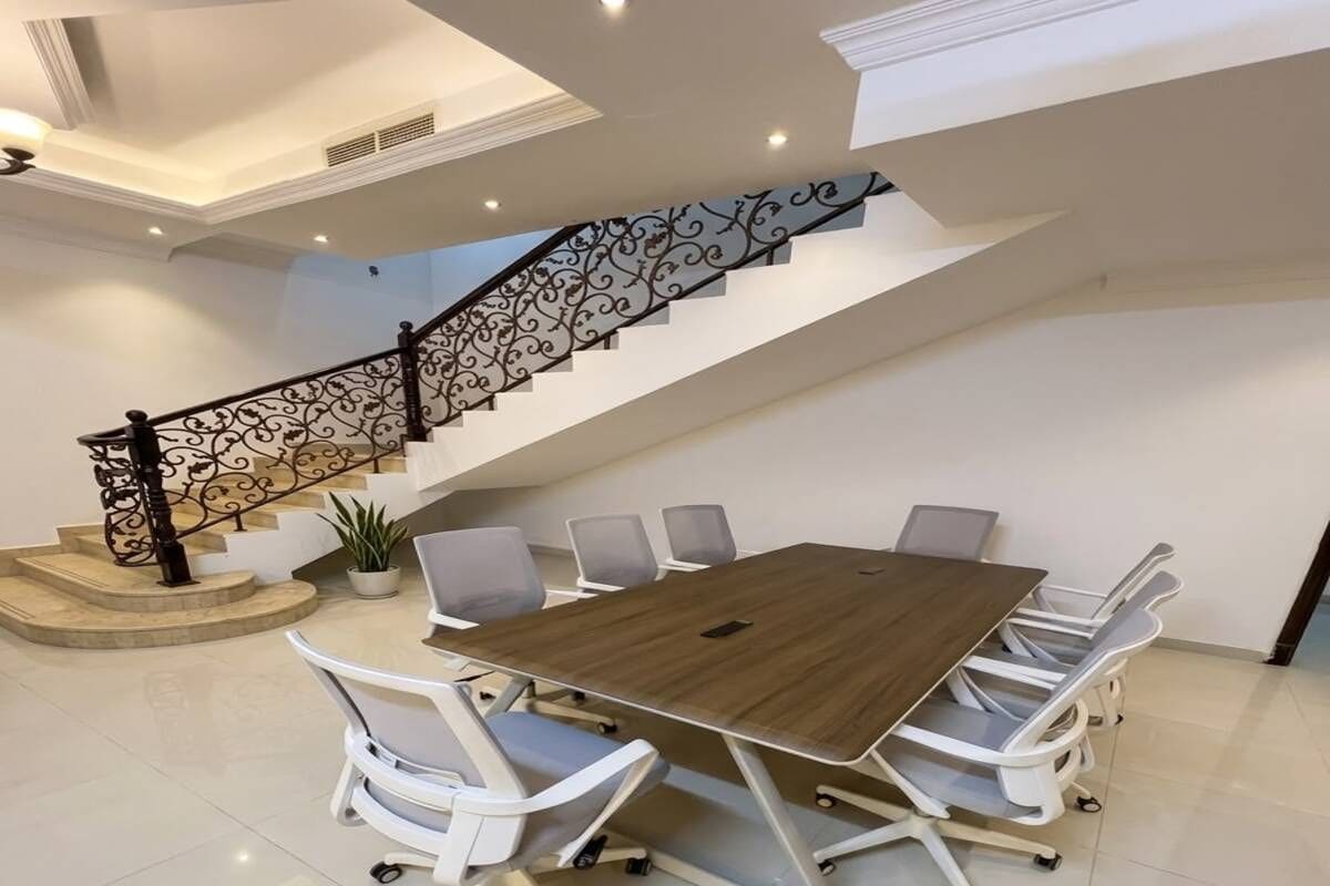 Un salón de conferencias con una gran mesa, sillas blancas y una escalera con barandales ornamentados.