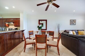 A modern kitchen with wooden cabinets, a dining area with a glass table and chairs, and a living area with a sofa and a ceiling fan. - Ocean-view Apt w/ Pool in Oceanica Resort (Cabo Velas)