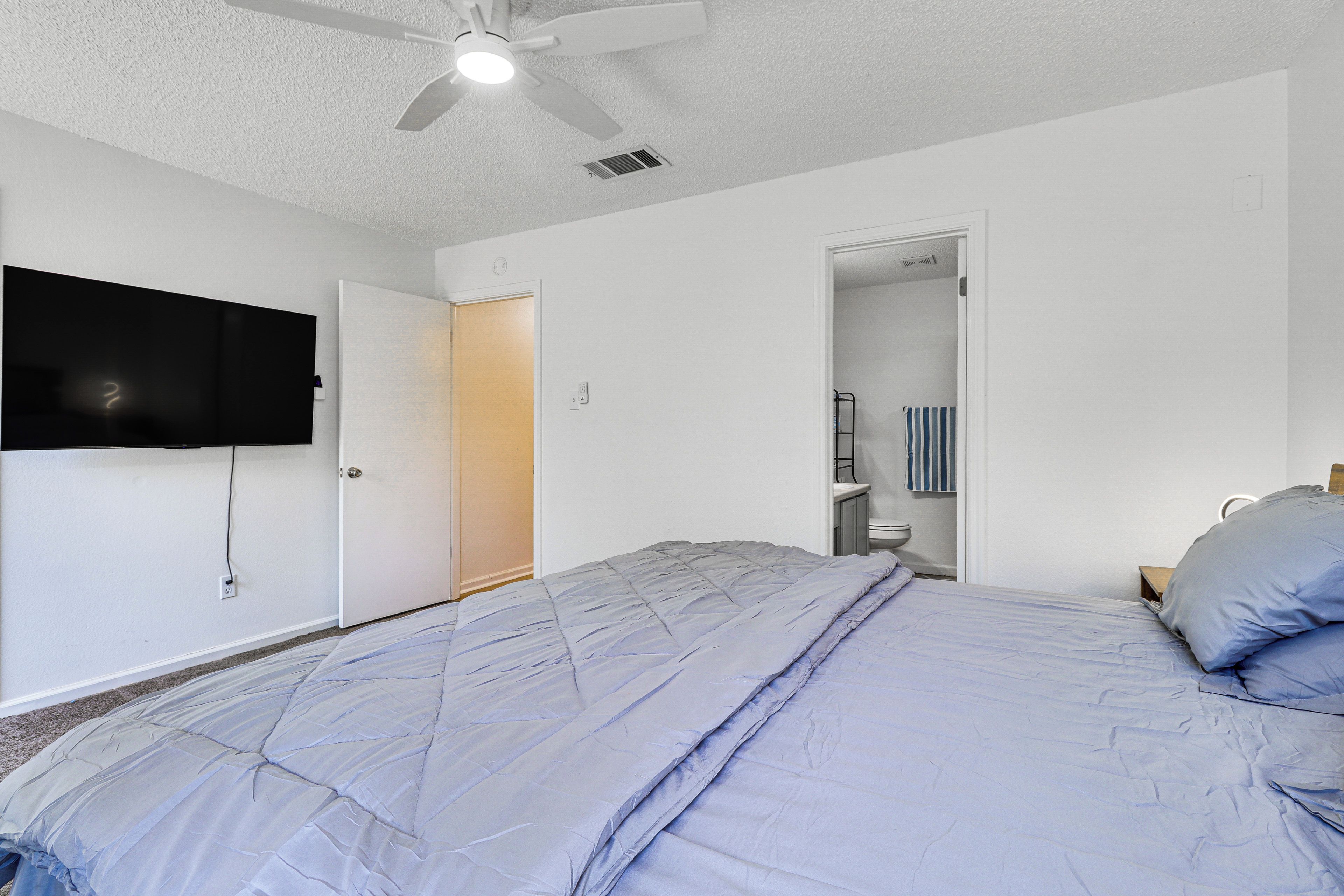 Un dormitorio con cama, ventilador de techo, televisor montado en la pared y puerta que da a un baño.