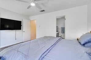 Un dormitorio con cama, ventilador de techo, televisor montado en la pared y puerta que da a un baño.