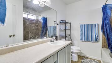 Un baño con lavamanos blanco, un espejo, inodoro, cortina de ducha y un perchero para toallas.