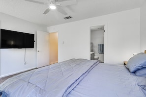 Un dormitorio con cama, ventilador de techo, televisor montado en la pared y puerta que da a un baño.