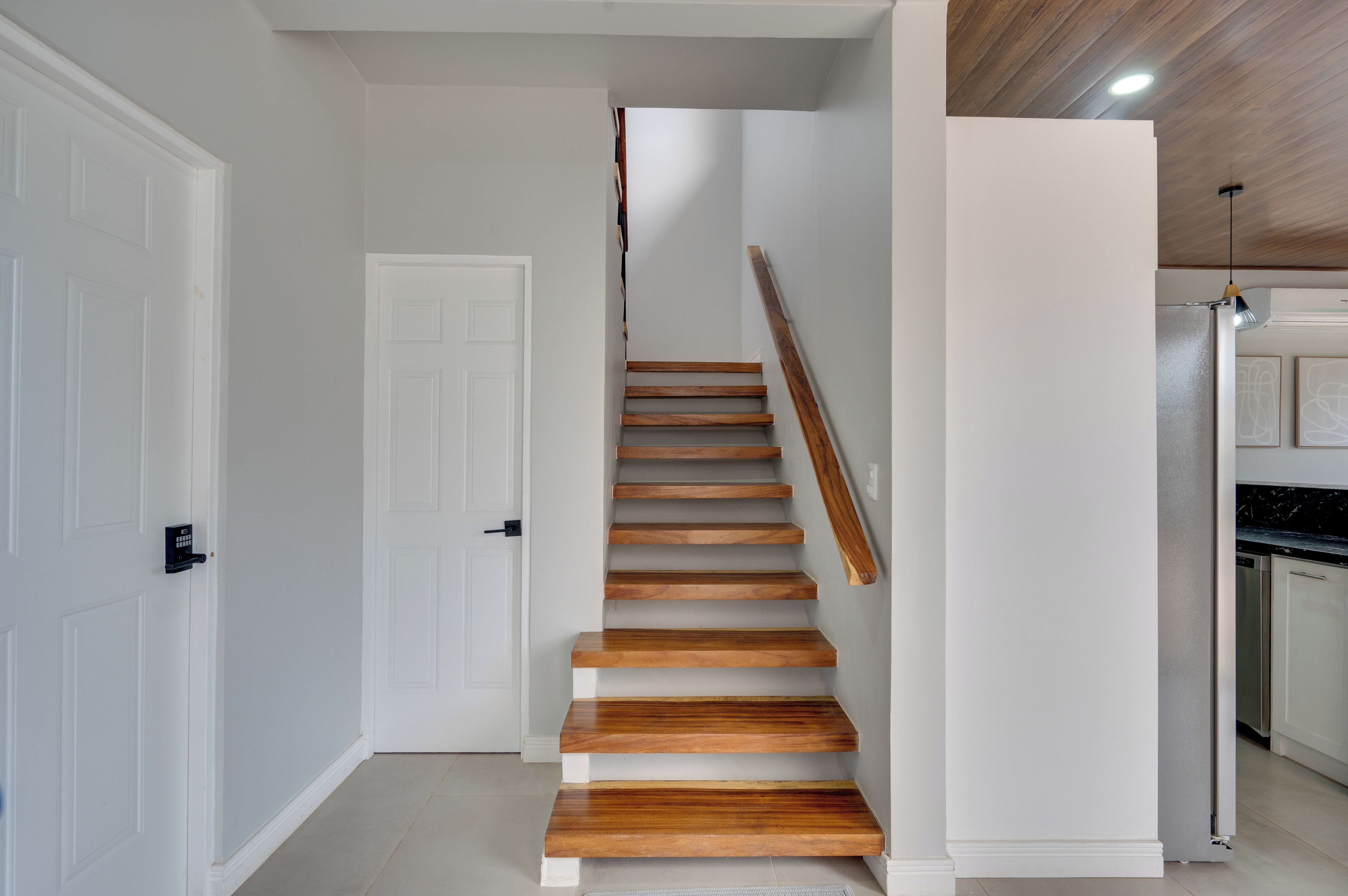 Una escalera de madera conduce a una plataforma, con paredes blancas y una zona de cocina que incluye un refrigerador.