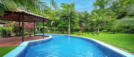 A swimming pool with a tiled edge, surrounded by lush greenery and a covered seating area.