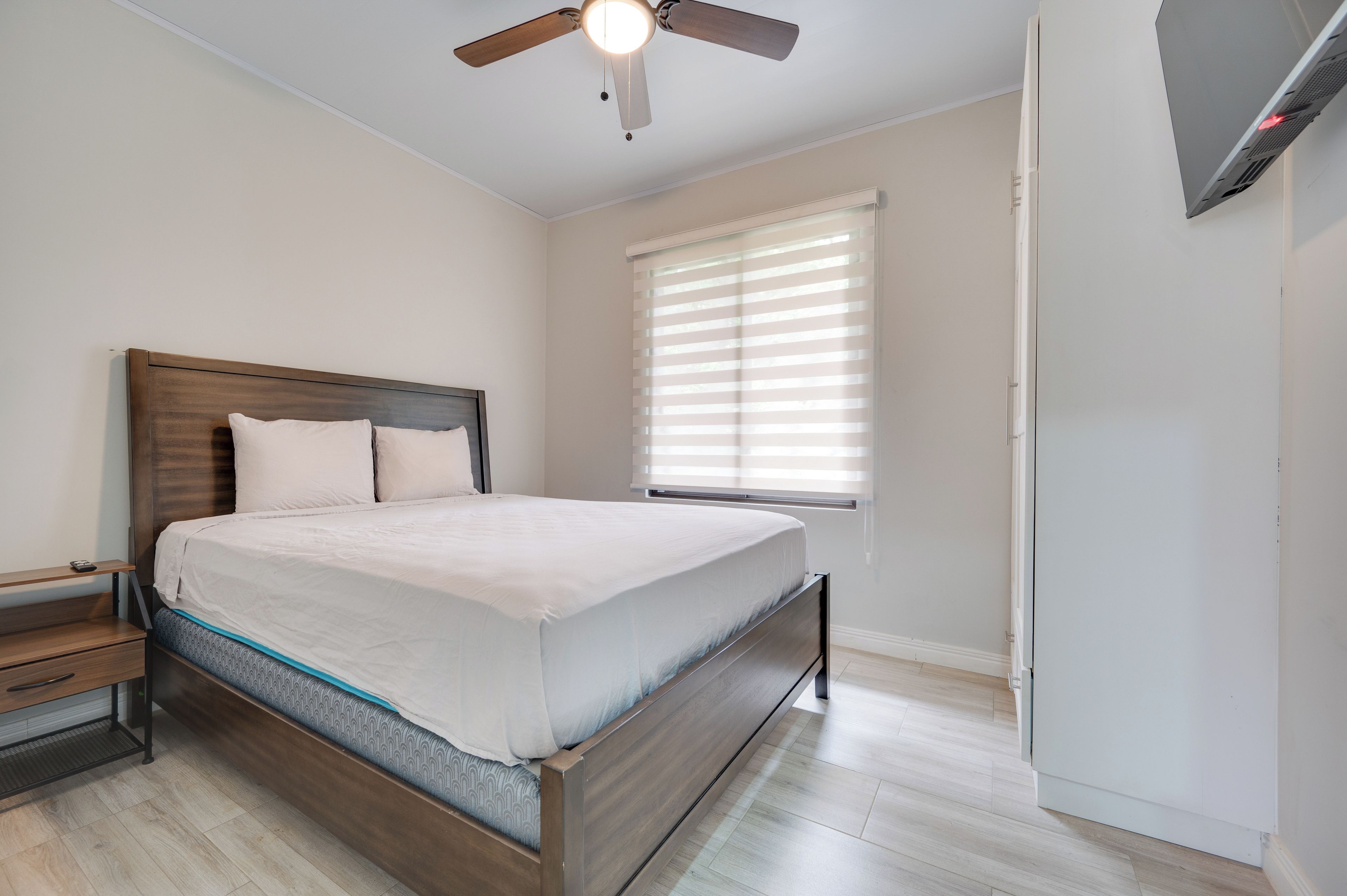 Un dormitorio con cama, mesita de noche, cabecera de madera, ventilador de techo y ventana con persianas.