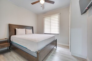 Quarto com cama, mesa de cabeceira, cabeceira de madeira, ventoinha no teto e janela com persianas.