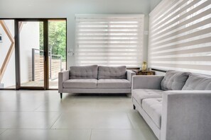 Un salon moderne avec un canapé gris, une porte-fenêtre coulissante donnant sur un espace extérieur et un mur équipé de stores horizontaux.