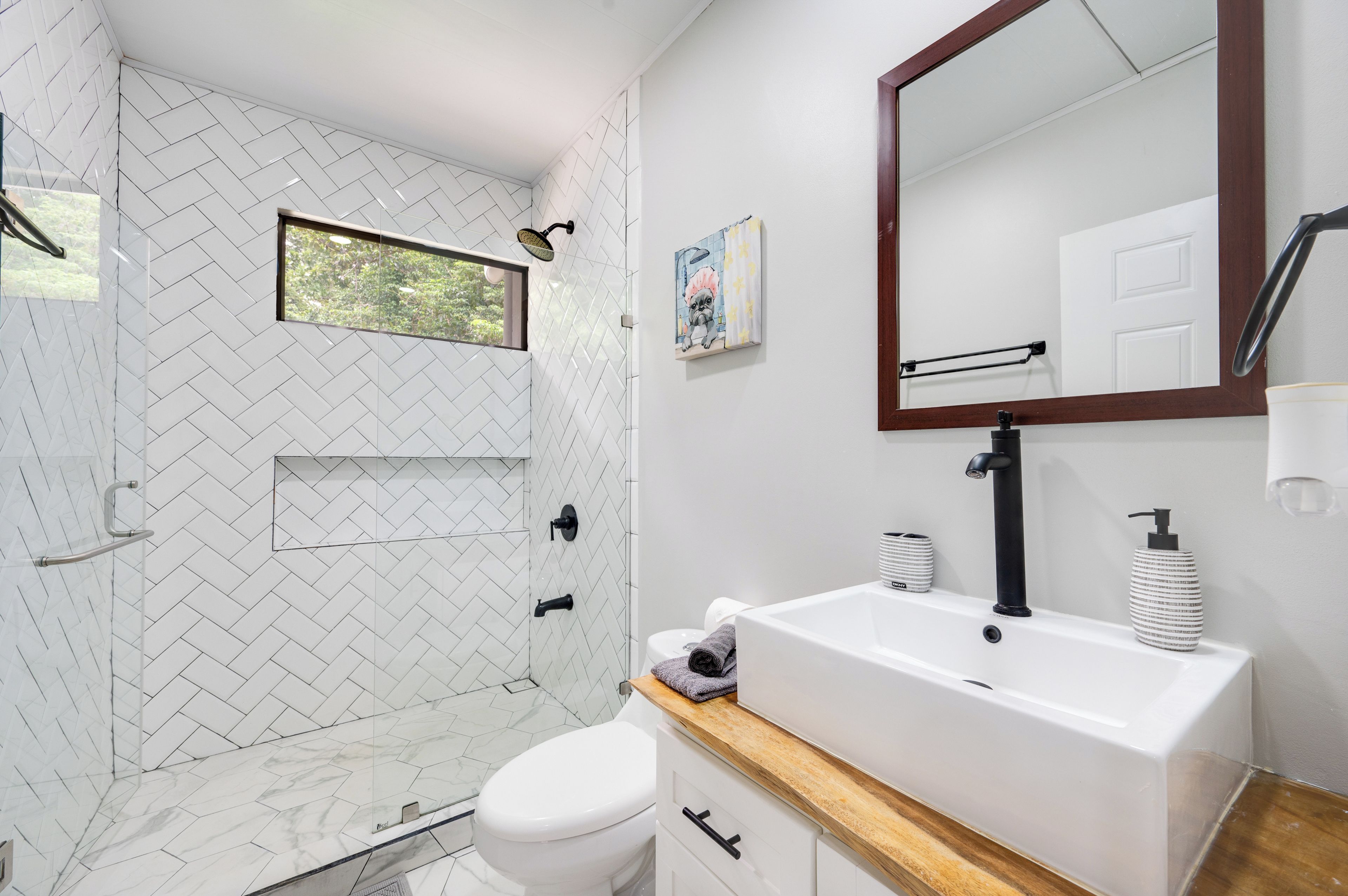 A modern bathroom with a herringbone tile pattern, a shower with a window, a white sink with a soap dispenser, and a mirror with a wooden frame.