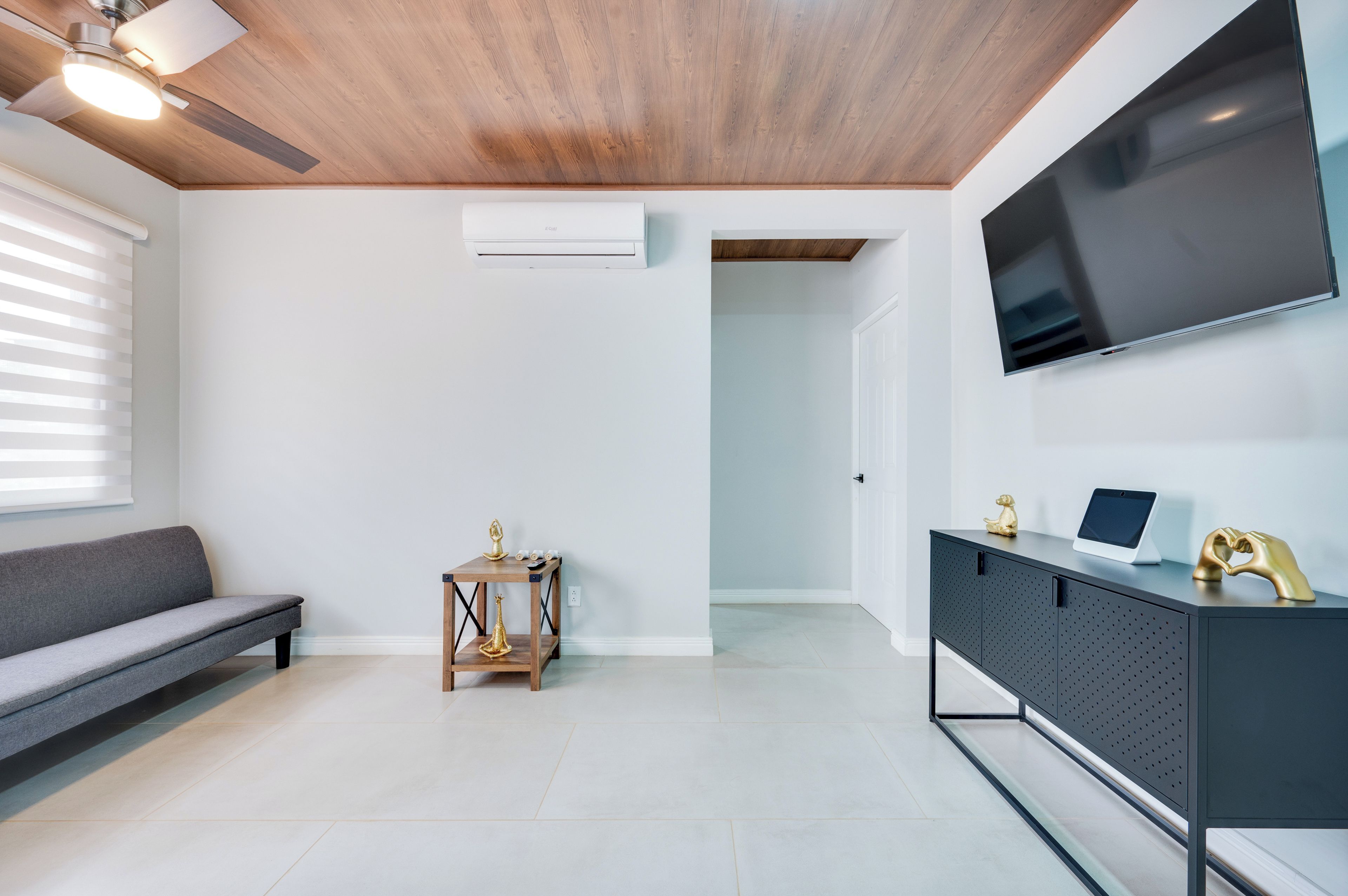 Una sala moderna con un sofá gris, una mesa de centro de madera, un televisor sobre un mueble negro y un ventilador de techo.