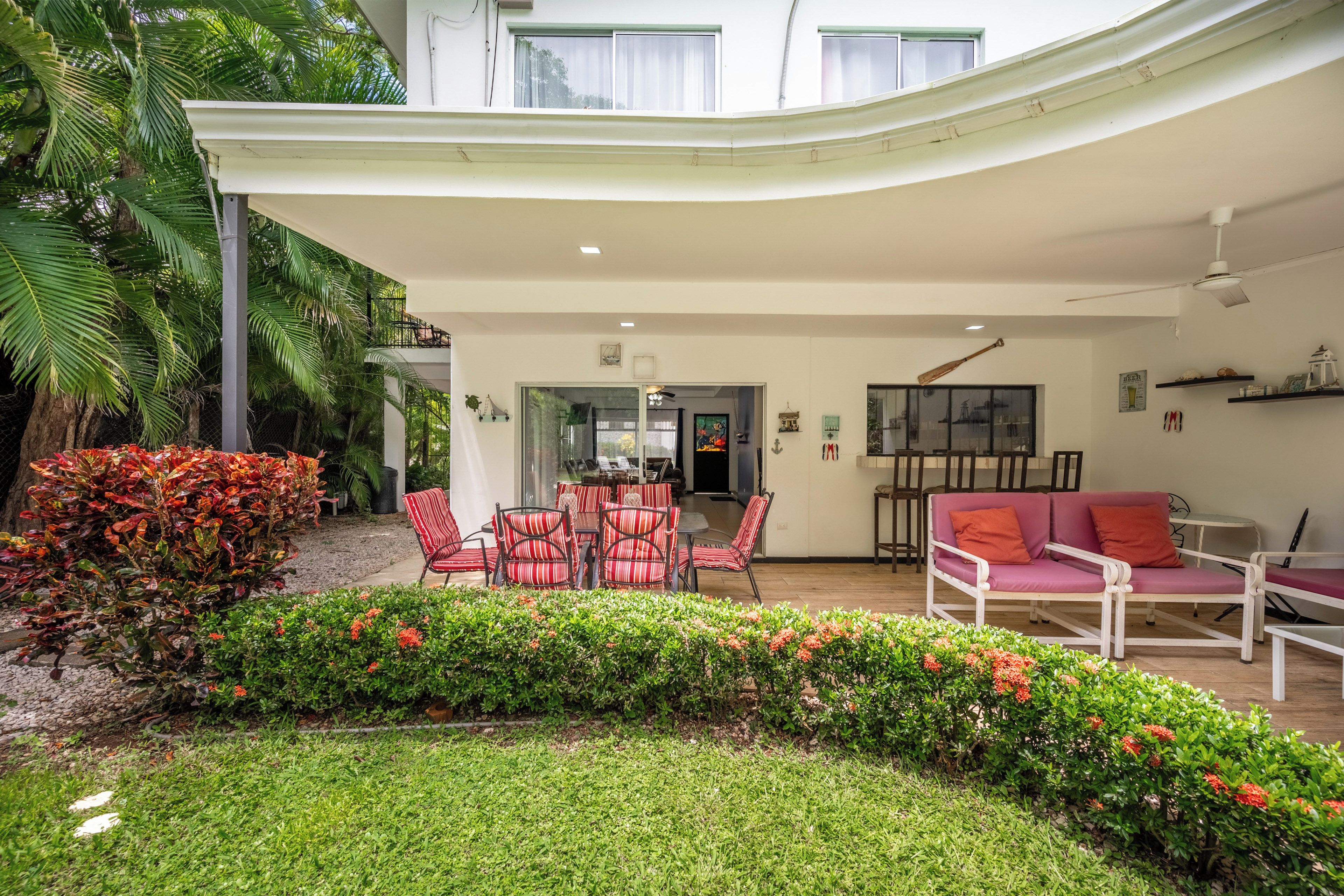 A covered outdoor seating area with chairs and tables, surrounded by greenery and a view of the interior.