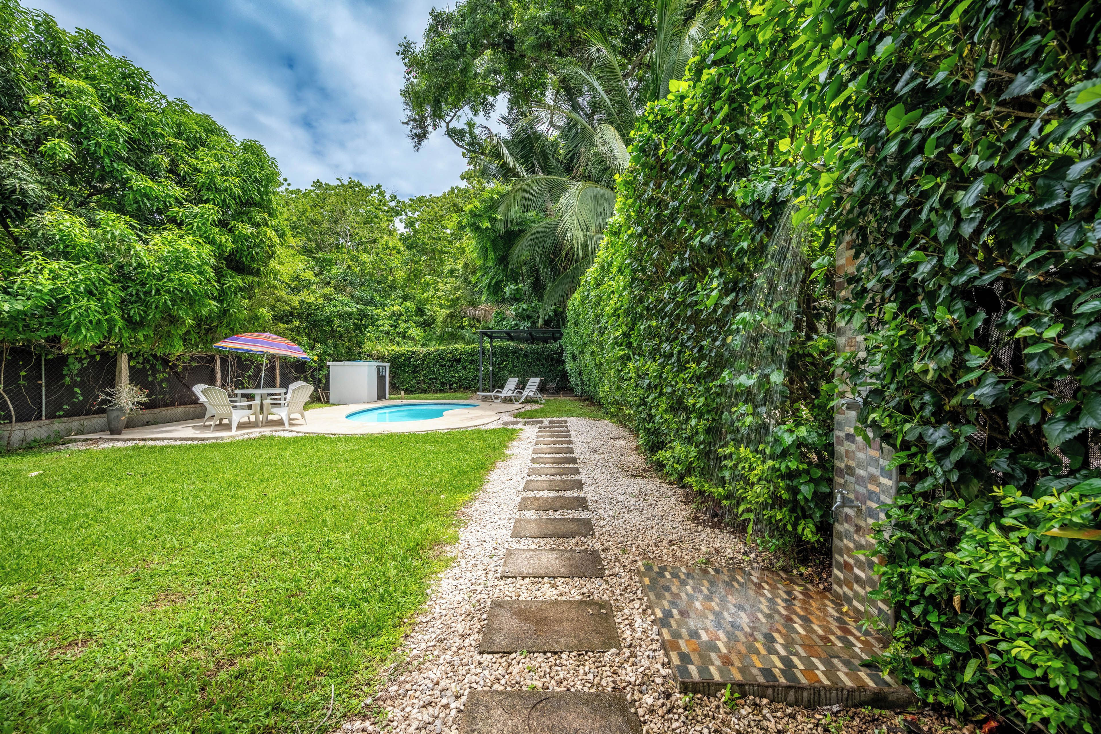 A backyard with a swimming pool, a patio with chairs, and a stone pathway leading to a door.