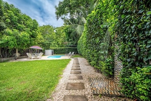 A backyard with a swimming pool, a patio with chairs, and a stone pathway leading to a door.