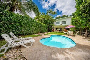 A swimming pool with lounge chairs, a covered patio with chairs, and a house with a green roof.