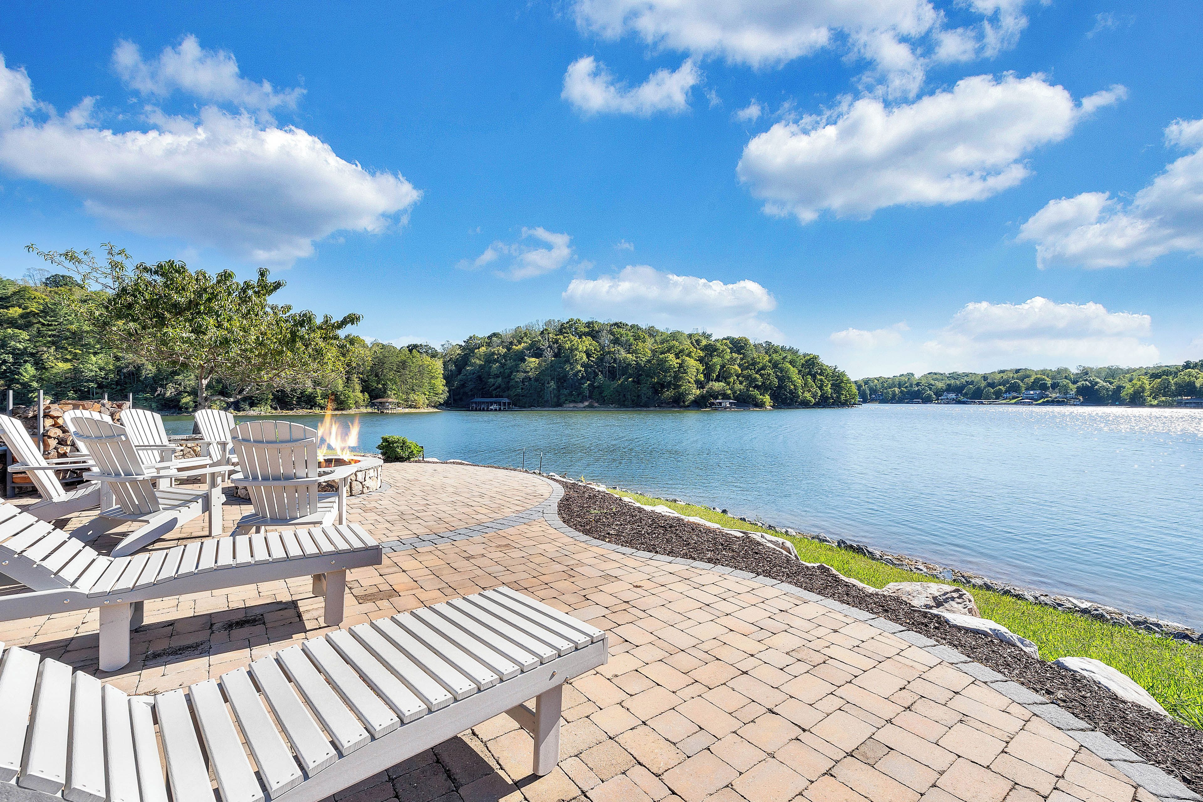A lakeside deck with Adirondack chairs and a fire pit, overlooking a calm lake and forested hills.