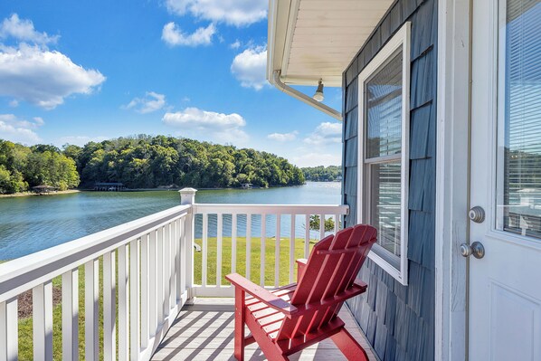 A wooden deck with a red Adirondack chair, overlooking a lake with a forested shore.