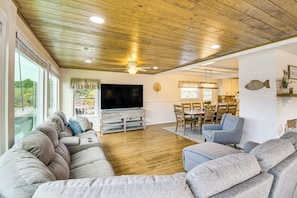 A spacious living room with a wooden ceiling, a flat-screen TV, a dining area with a table and chairs, and a view through large windows.