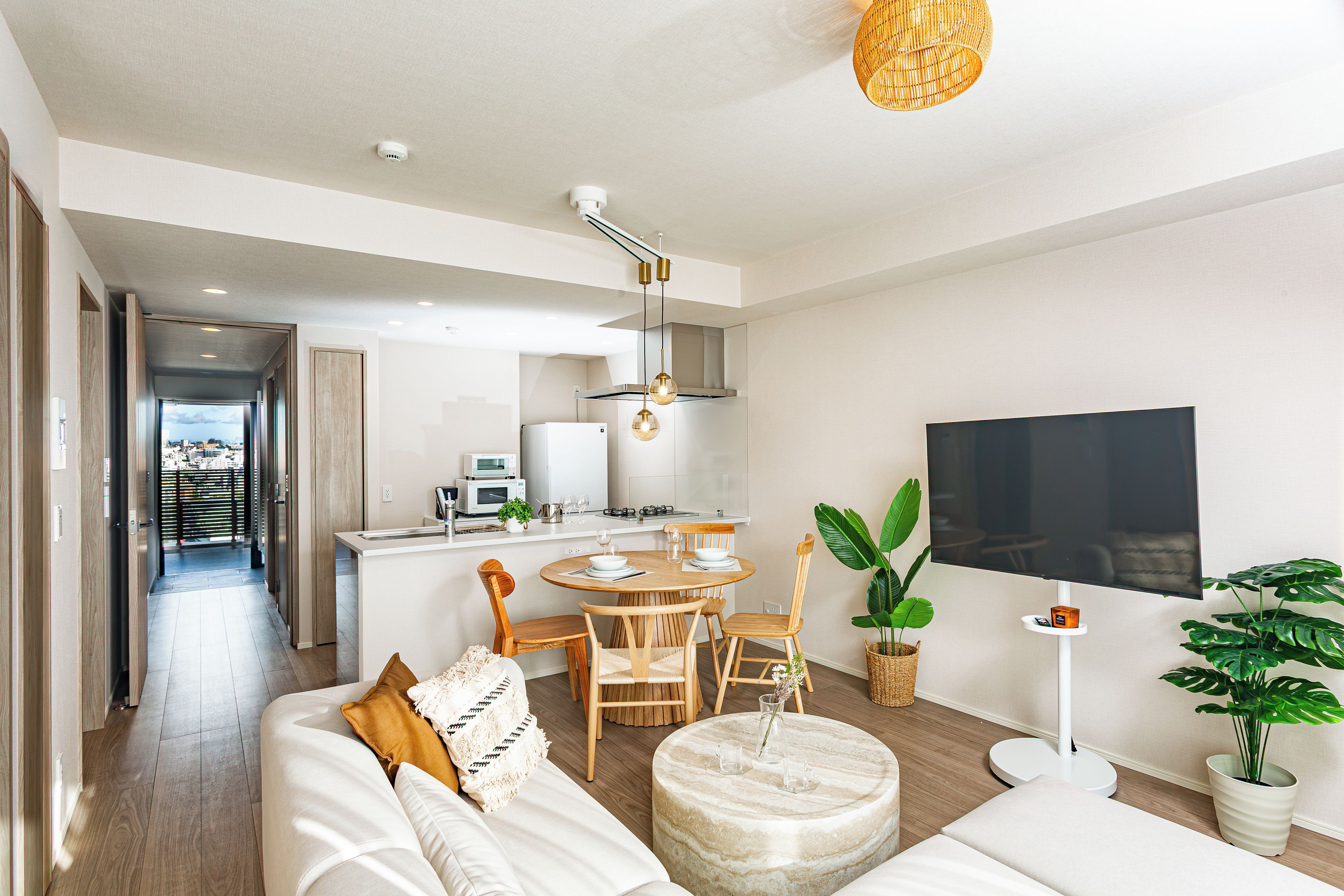 A modern living room with a sofa, dining table, and chairs, a kitchen area with appliances, and a TV mounted on the wall.