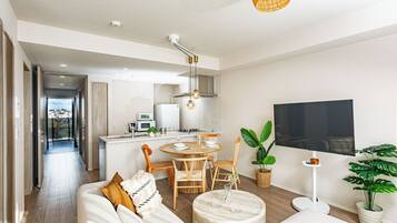 A modern living room with a sofa, dining table, and chairs, a kitchen area with appliances, and a TV mounted on the wall.