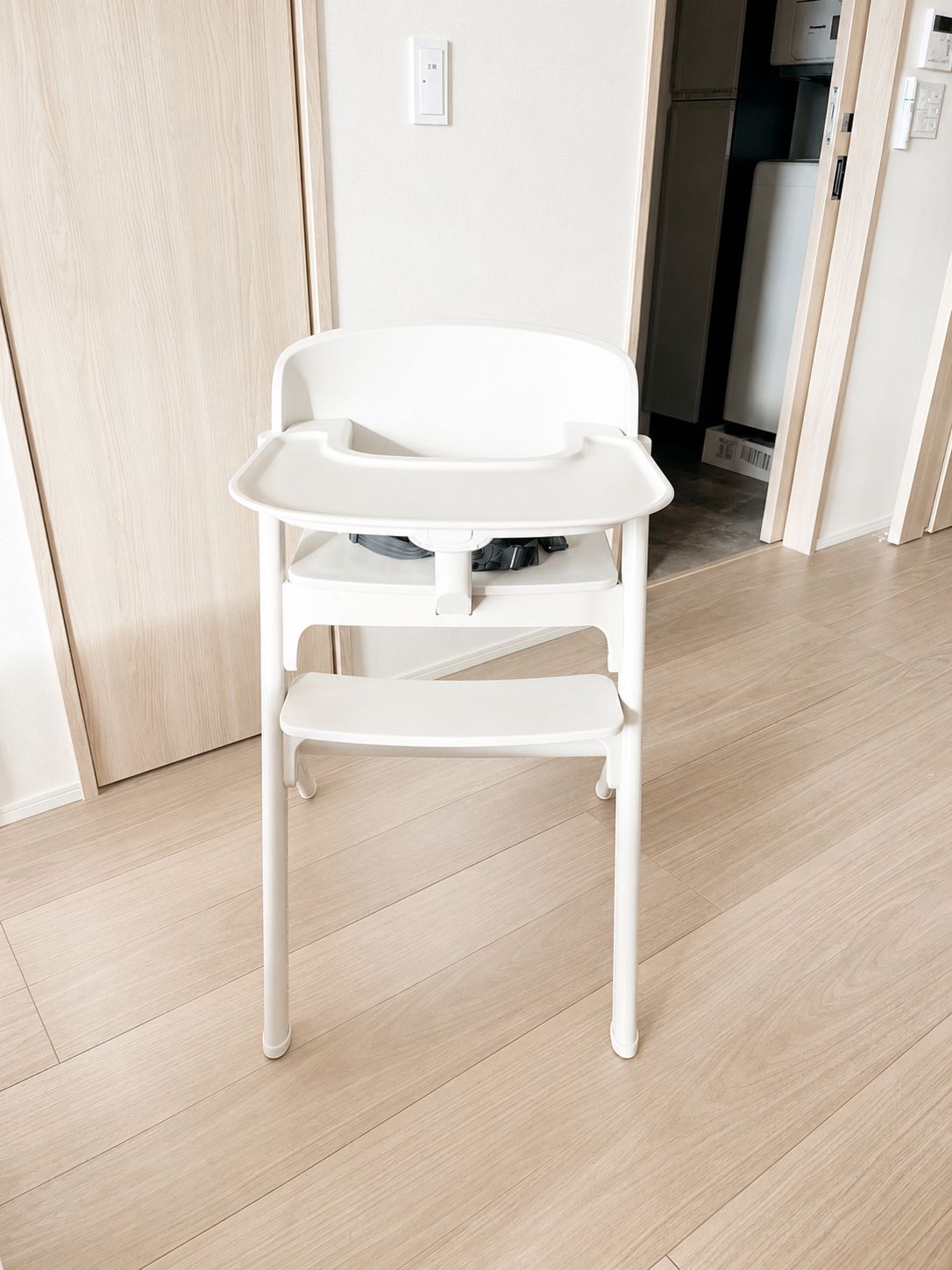A white high chair with a tray table in a room with wooden flooring and a door in the background.