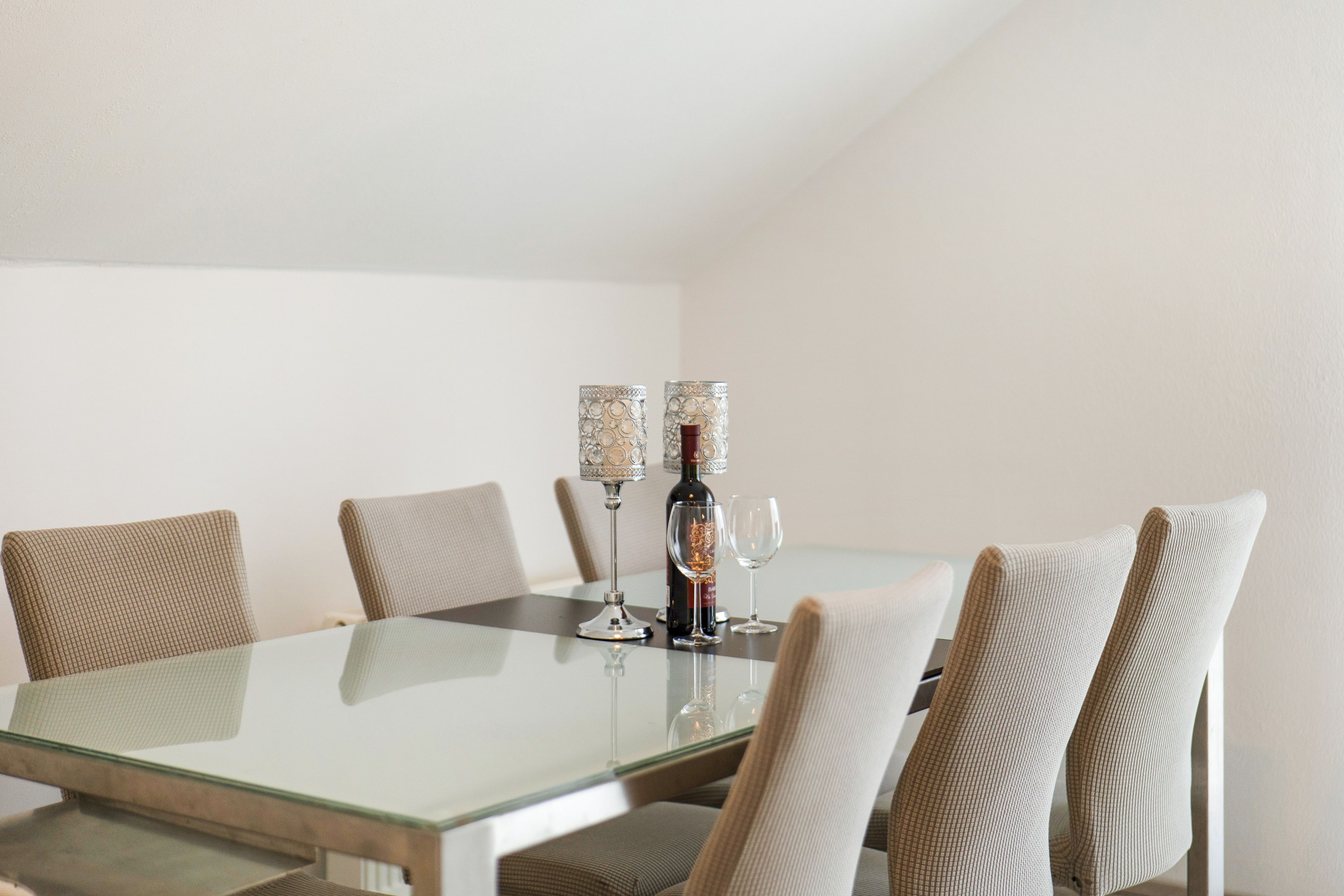 Un comedor con una mesa de vidrio, seis sillas, dos candelabros, una botella de vino y dos copas de vino.