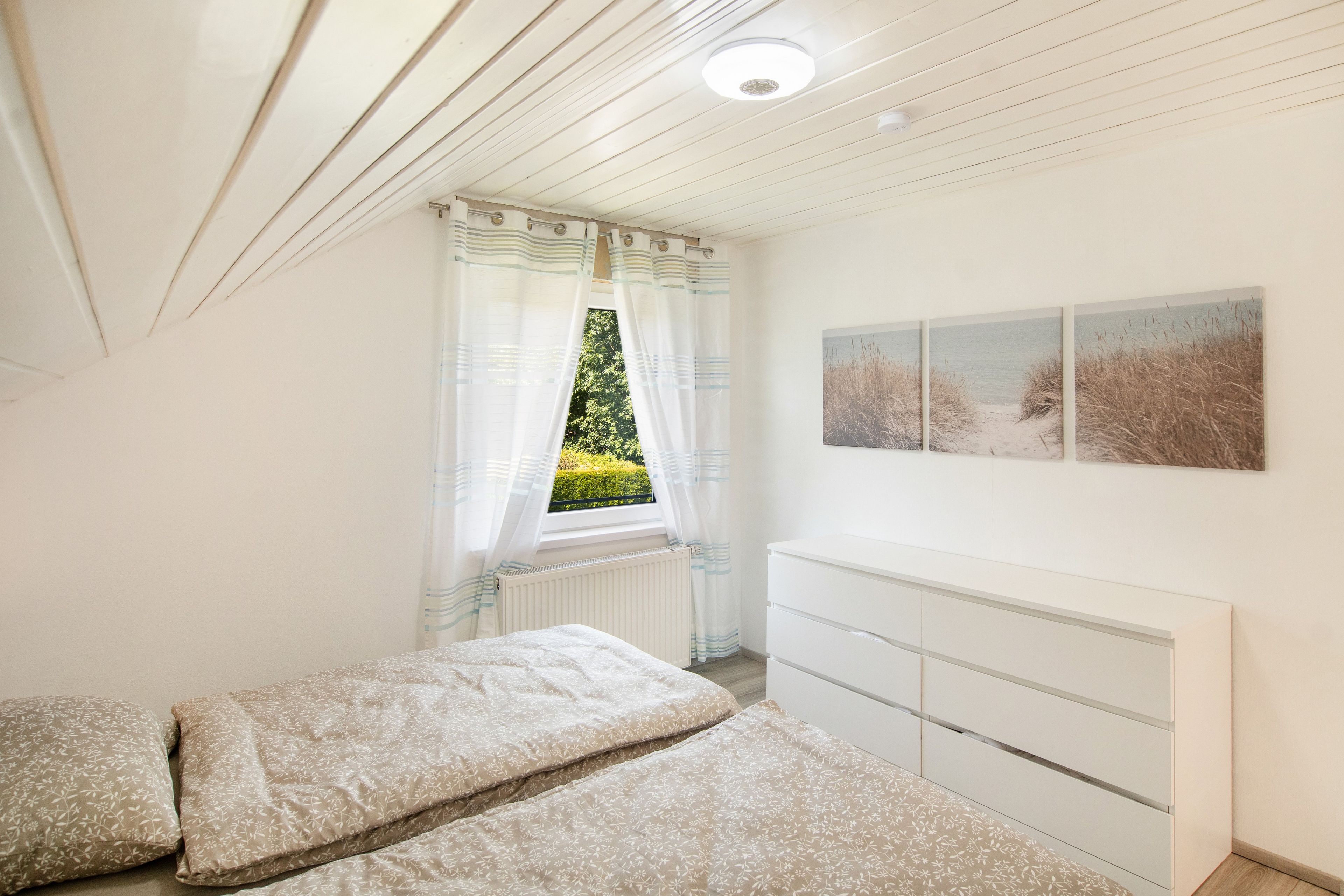 Een slaapkamer met een bed, een dressoir, een raam met gordijnen en een schilderij van een strand.
