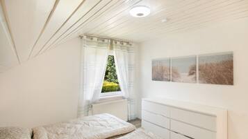 Een slaapkamer met een bed, een dressoir, een raam met gordijnen en een schilderij van een strand.