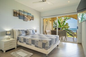 A bedroom with a bed, bedside tables, a nightstand, a view of the ocean through a sliding door, and a ceiling fan. - Oceanfront 3BR Condo in Playa Potrero (Tempate)