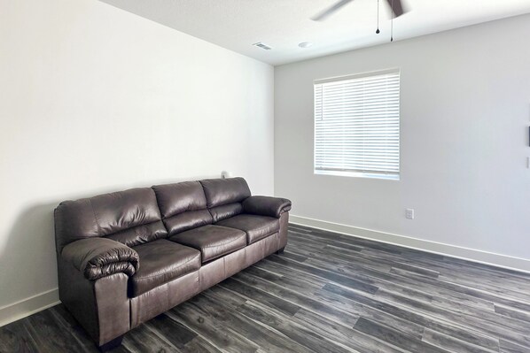 A room with a dark brown leather sofa, a window with blinds, and a ceiling fan. - Walk to Parks & Dining! Newly Built Wichita Condo (Wichita)