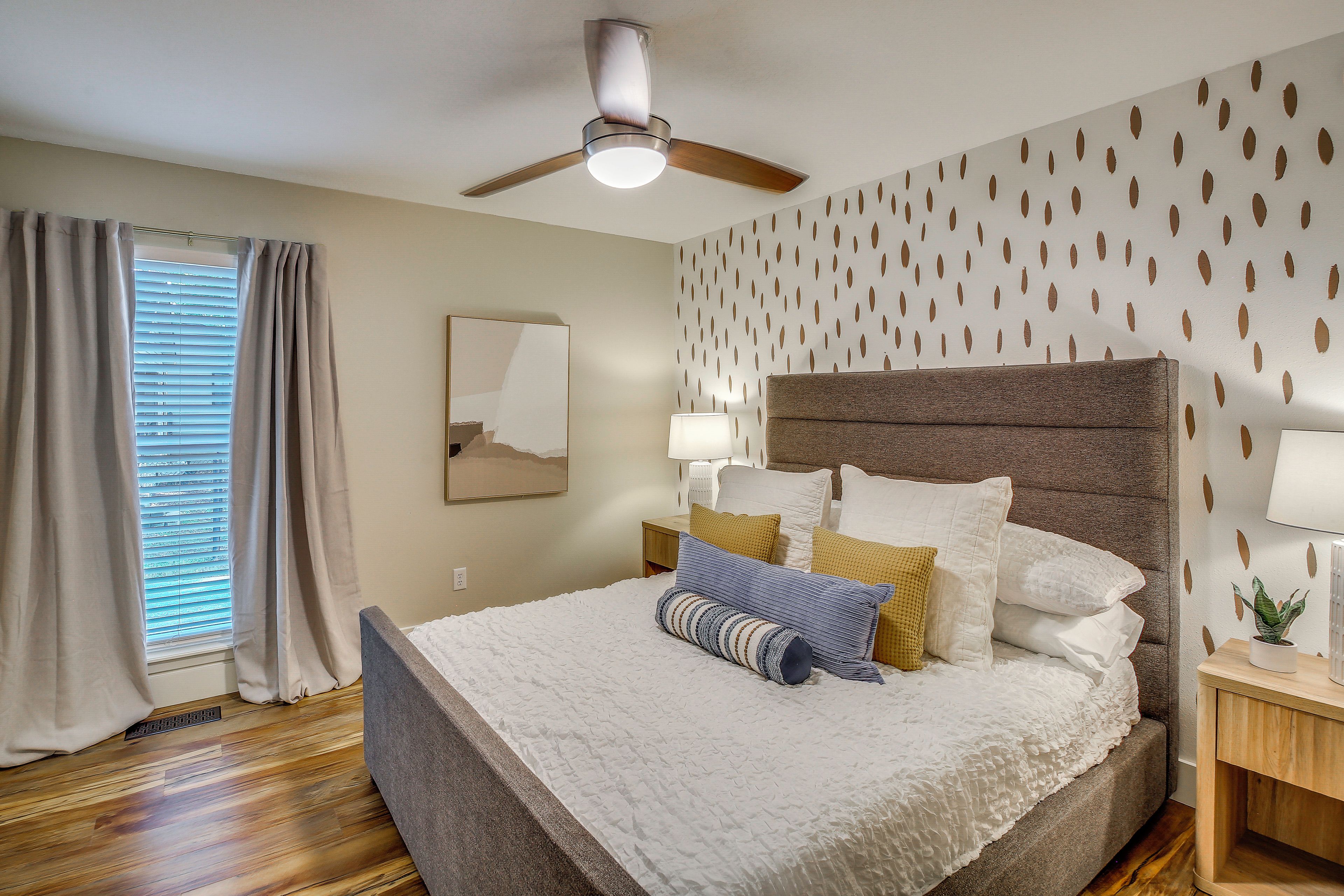 Une chambre à coucher comprenant un lit, un ventilateur de plafond, une table de chevet, une lampe, une fenêtre avec des rideaux et un mur orné d’un motif de feuilles.