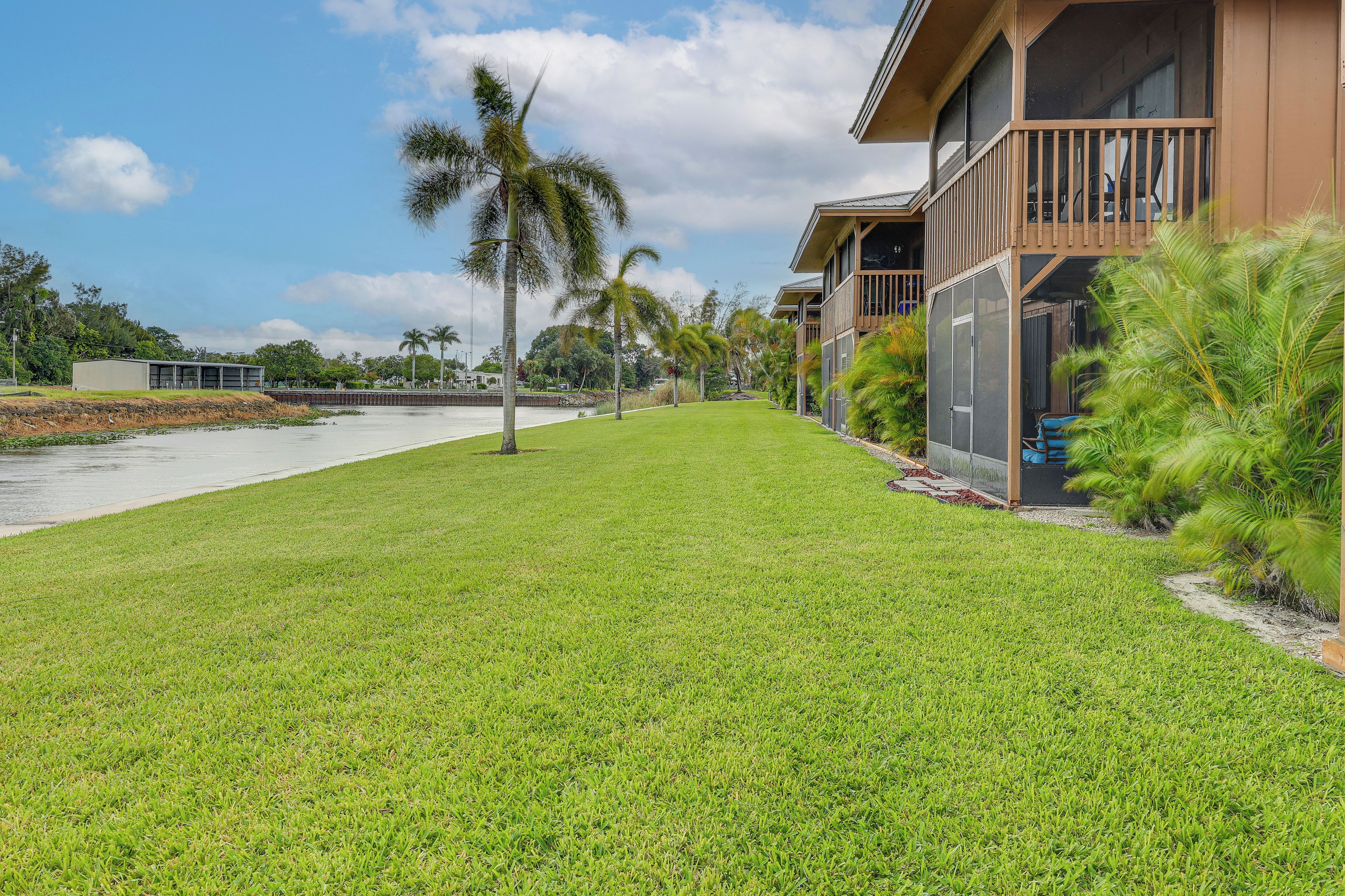 A well-maintained grassy area with a water body, palm trees, and a building with a balcony.