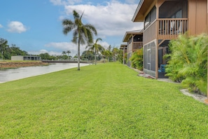 A well-maintained grassy area with a water body, palm trees, and a building with a balcony. - Lake Okeechobee Condo w/ Pool & Patio! (Clewiston)
