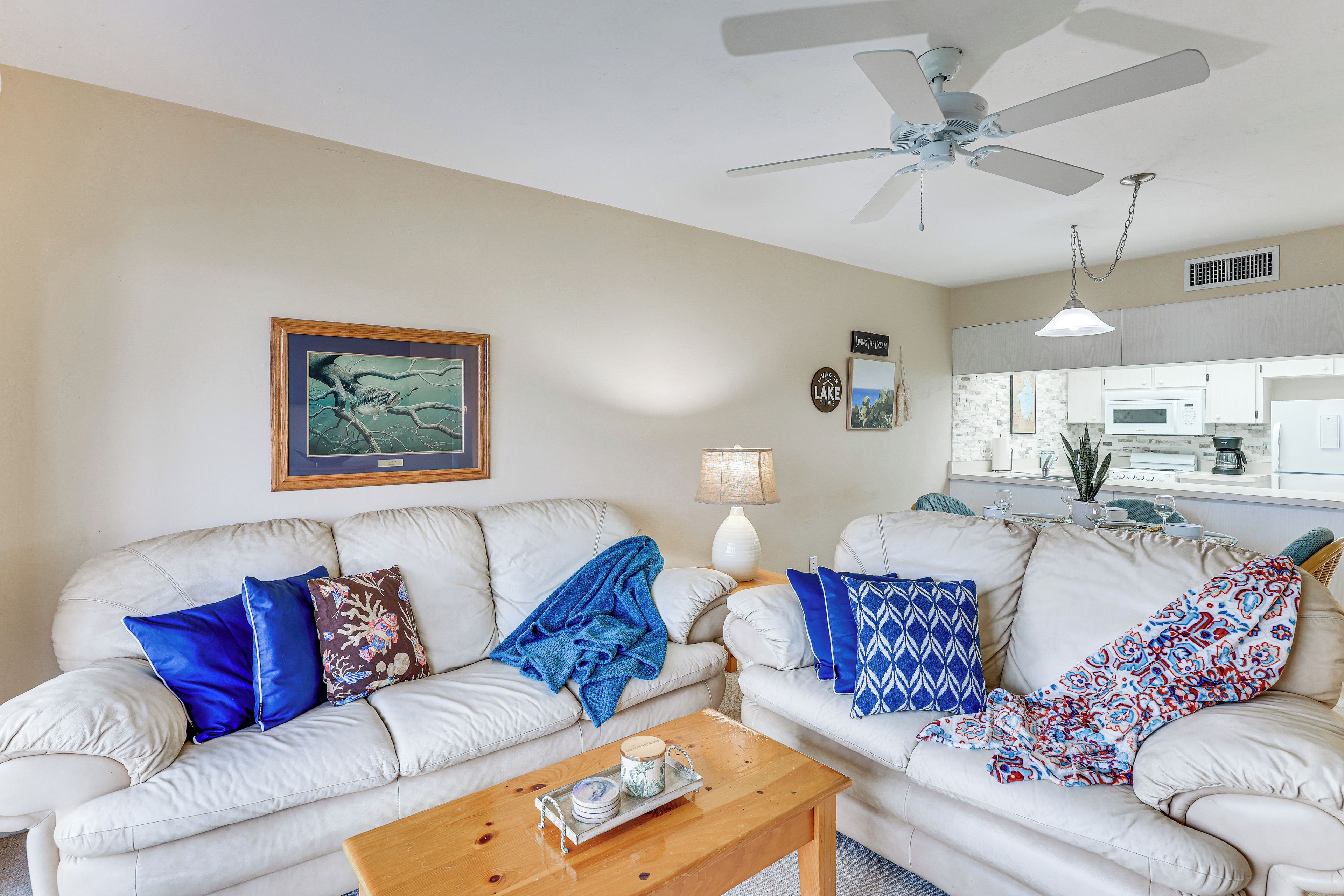 A living room with two white sofas, a wooden coffee table, a ceiling fan, and a kitchen visible through an open door.