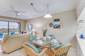 A dining area with a glass table, wicker chairs, and a view of a building through large windows. - Lake Okeechobee Condo w/ Pool & Patio! (Clewiston)