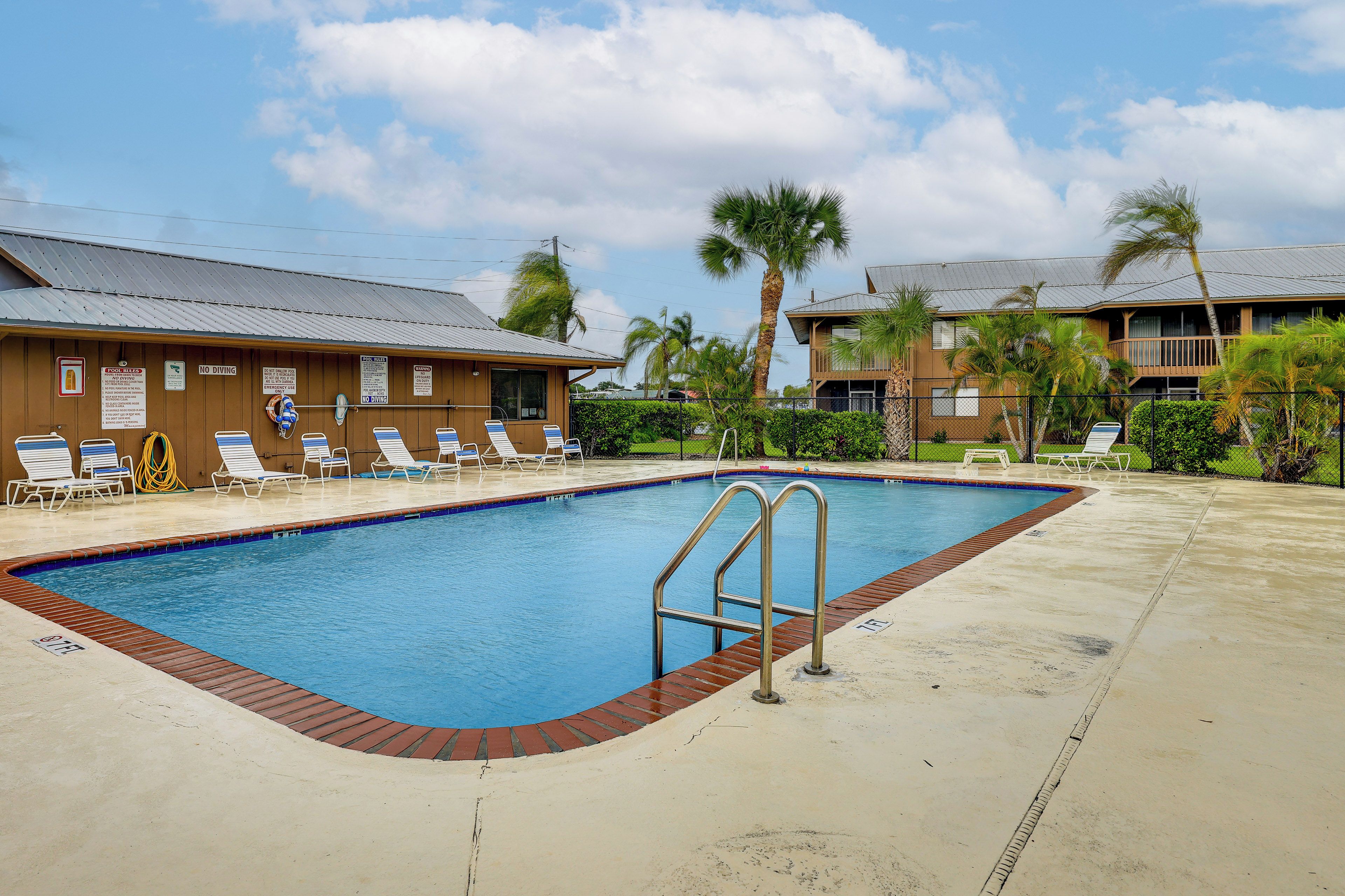A swimming pool with a metal ladder, surrounded by lounge chairs and palm trees, with a building in the background.