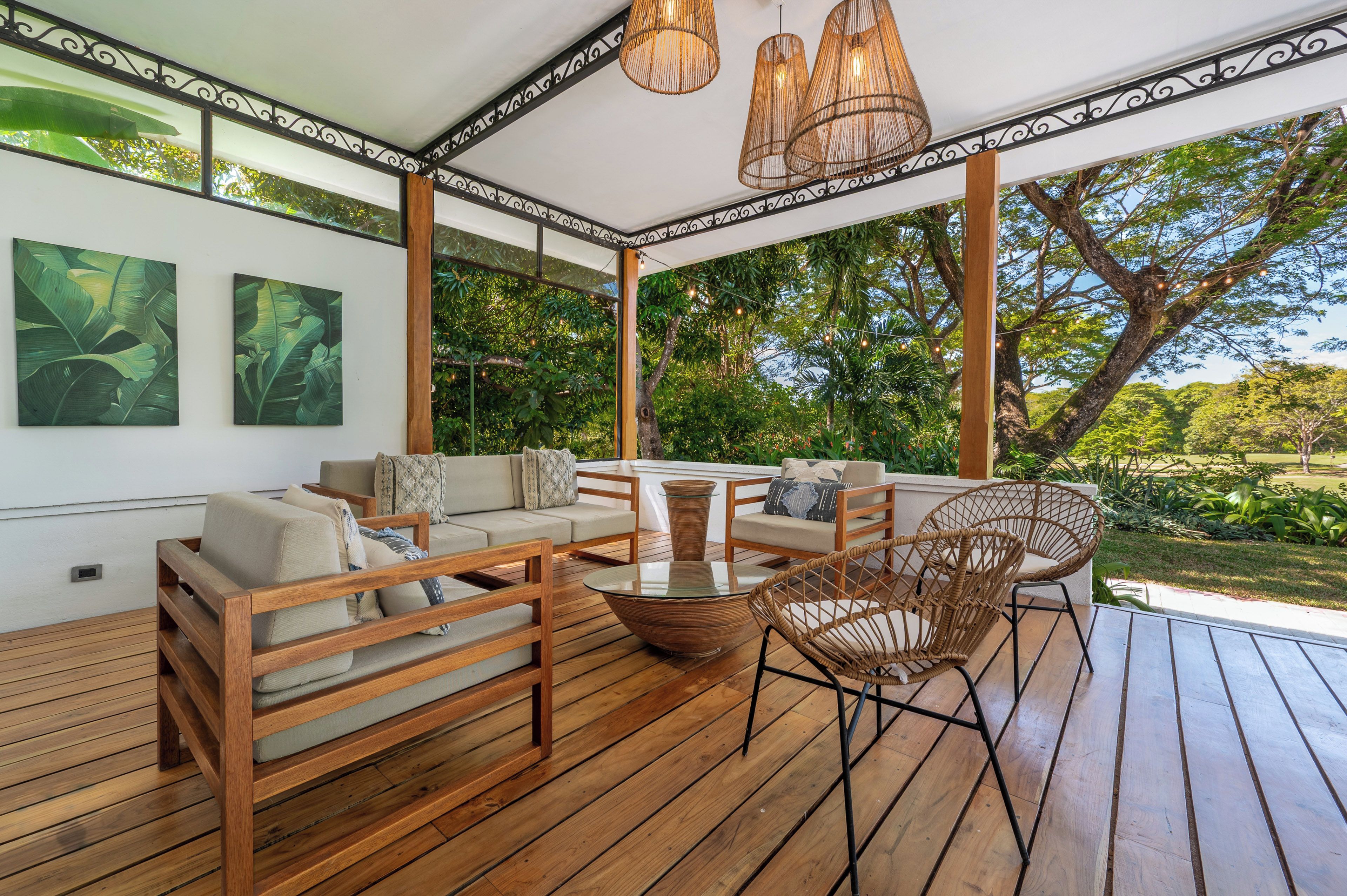 A covered outdoor seating area with wooden furniture, a glass table, and hanging wicker lamps.