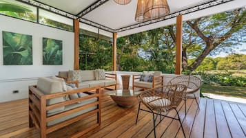 A covered outdoor seating area with wooden furniture, a glass table, and hanging wicker lamps.