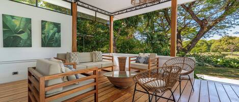 Un espacio de estar al aire libre cubierto con muebles de madera, una mesa de vidrio y lámparas colgantes de mimbre.