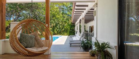 Une terrasse en bois avec un fauteuil suspendu, une piscine et des plantes en pot.