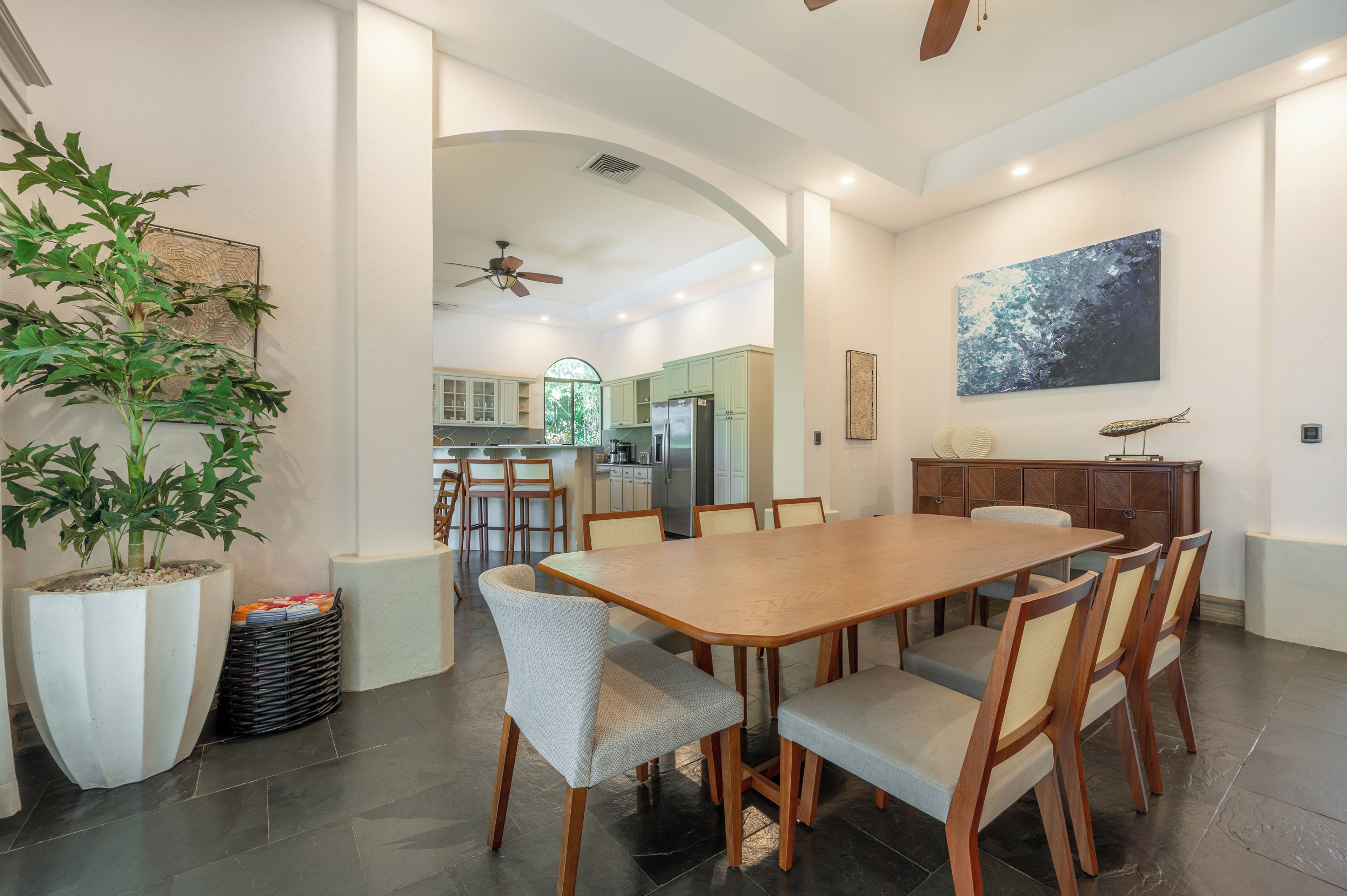 A spacious dining area with a large wooden table and chairs, a ceiling fan, and a view into the kitchen area.