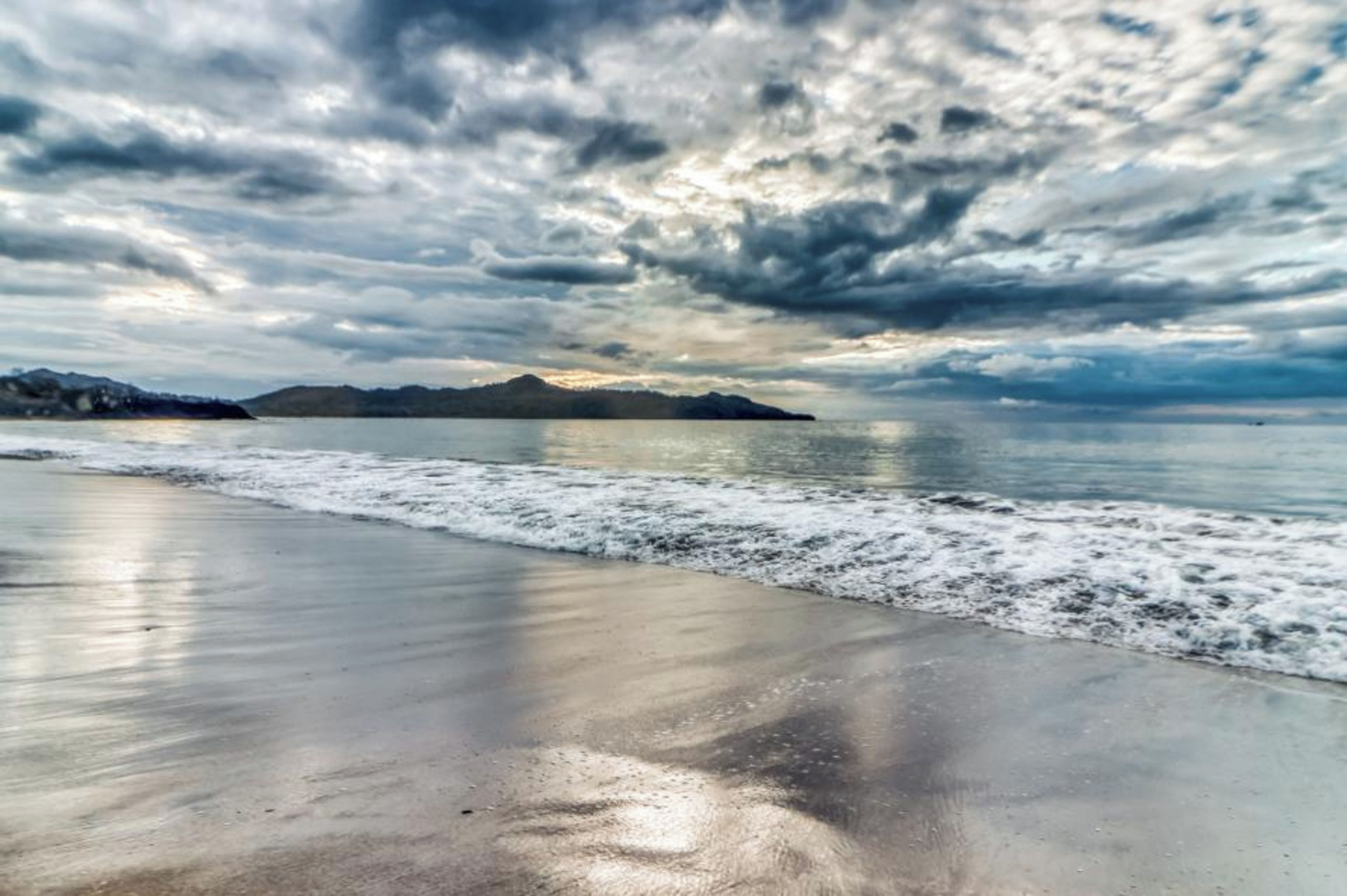 A beach with a calm sea, a wet sand foreground, and a cloudy sky.
