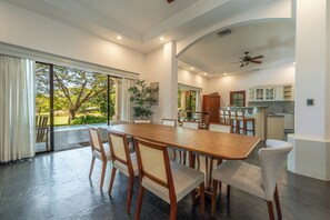 Un comedor amplio con una gran mesa y sillas de madera, una cocina con armarios, y vistas a un jardín a través de puertas correderas grandes.