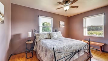 A bedroom with a bed, two windows, a ceiling fan, a bedside table, and a bench.