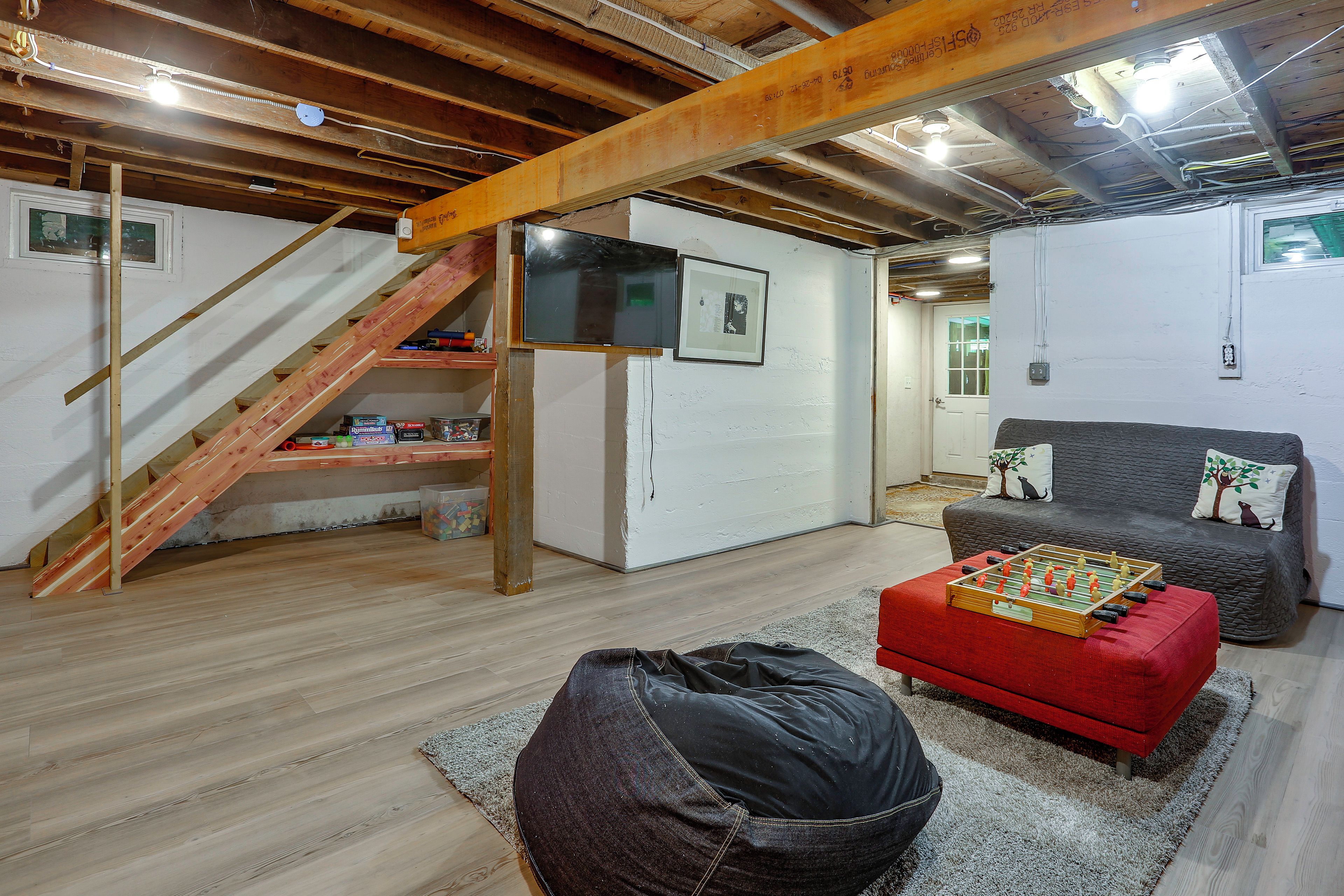 A room with a wooden staircase, a sofa, a red ottoman, and a foosball table.