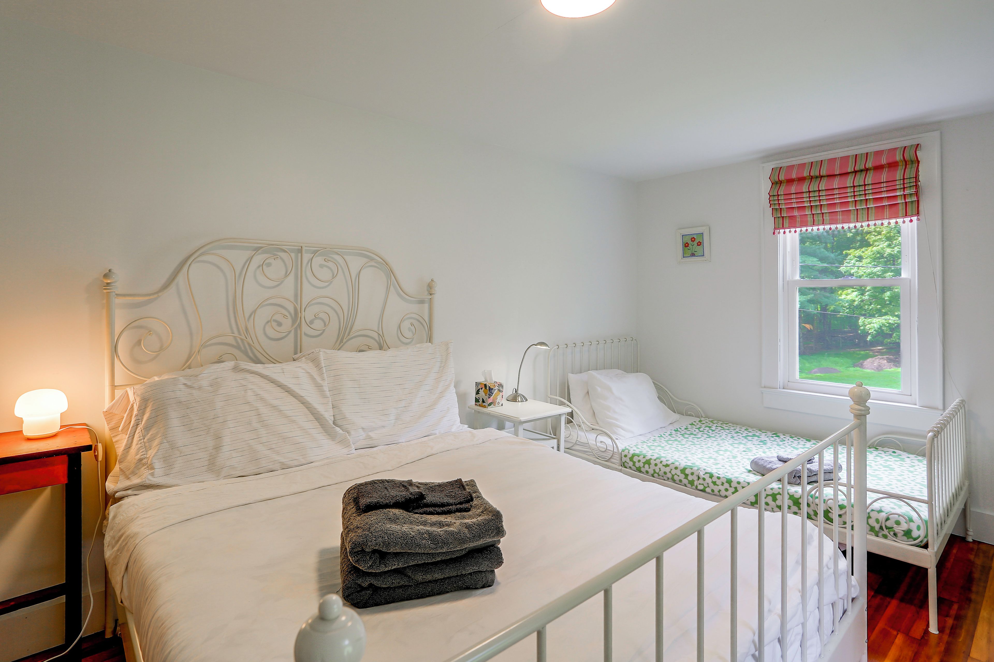 A bedroom with a double bed, a smaller bed, a nightstand with a lamp, and a window with a view of greenery.