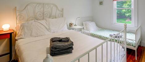 A bedroom with a double bed, a smaller bed, a nightstand with a lamp, and a window with a view of greenery.