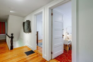 A hallway with wooden flooring, white walls, and a door leading to a bedroom with a bed and a nightstand.